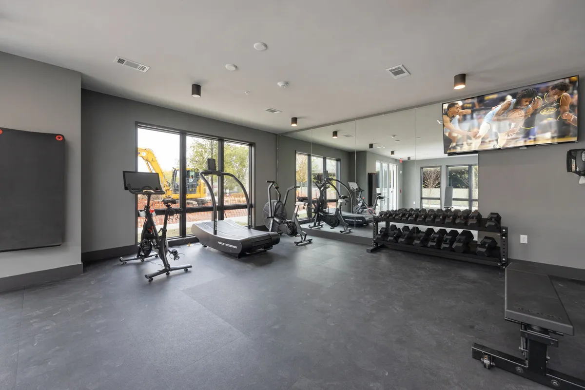 A well-equipped fitness center featuring a treadmill, free weights, exercise bike, large mirrors, and a TV. The space is bright, modern, and designed for both solo and group workouts, promoting wellness and energy.