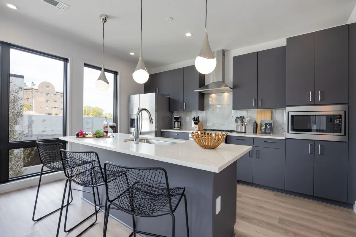 A bright, contemporary kitchen featuring a spacious island with bar stools, sleek pendant lights, and large windows that fill the space with natural light. The open-concept design encourages both cooking and socializing, while the backdrop includes tasteful décor and modern appliances.