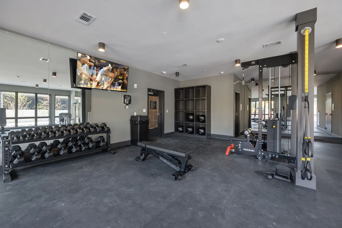 An interior view of the modern fitness center, featuring a dumbbell rack, weight bench, multifunctional gym equipment, and a large television screen. The space is bright and designed for group workouts or solo training.