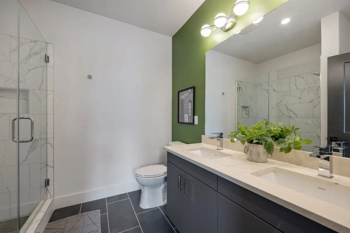 A modern bathroom featuring a double sink vanity with ample counter space, a glass shower enclosure, a vibrant green accent wall, and a potted plant adding a touch of freshness. The ambiance is calm and upscale.