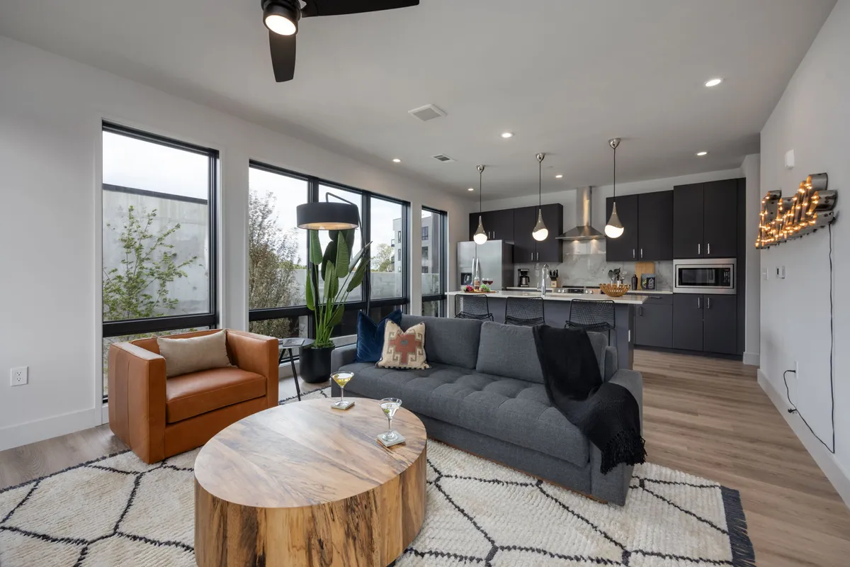 A spacious open-concept living room featuring a plush sofa, a stylish coffee table, and potted plants, with the kitchen area visible in the background. The space is filled with natural light from large windows, creating a welcoming atmosphere perfect for socializing or relaxing.