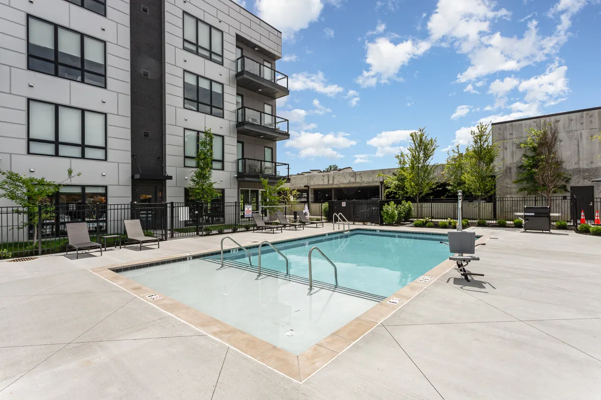 Exterior view of the sparkling swimming pool with lounge chairs, surrounding greenery, and the modern building facade in the background. The tranquil setting suggests leisurely afternoons and community events.