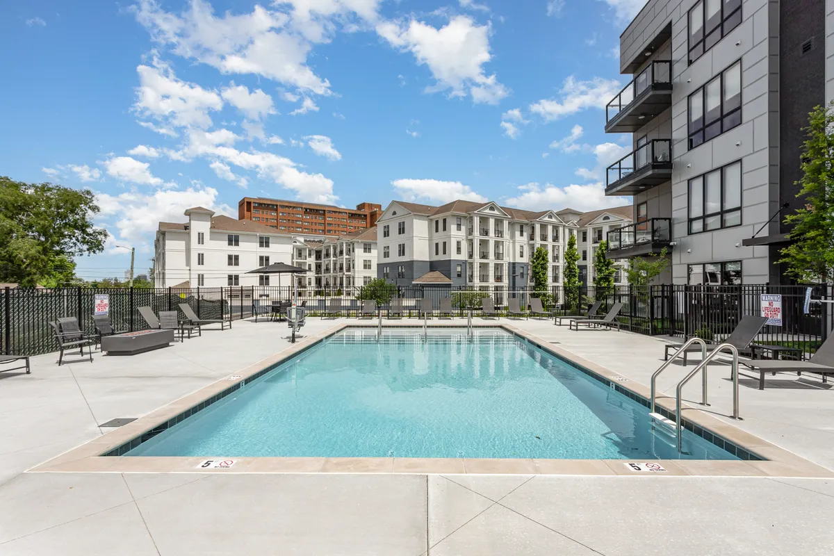 An inviting outdoor swimming pool surrounded by modern apartment buildings and a comfortable lounge area. The scene is bathed in natural light, with seating for relaxing or socializing and ample space for group activities.