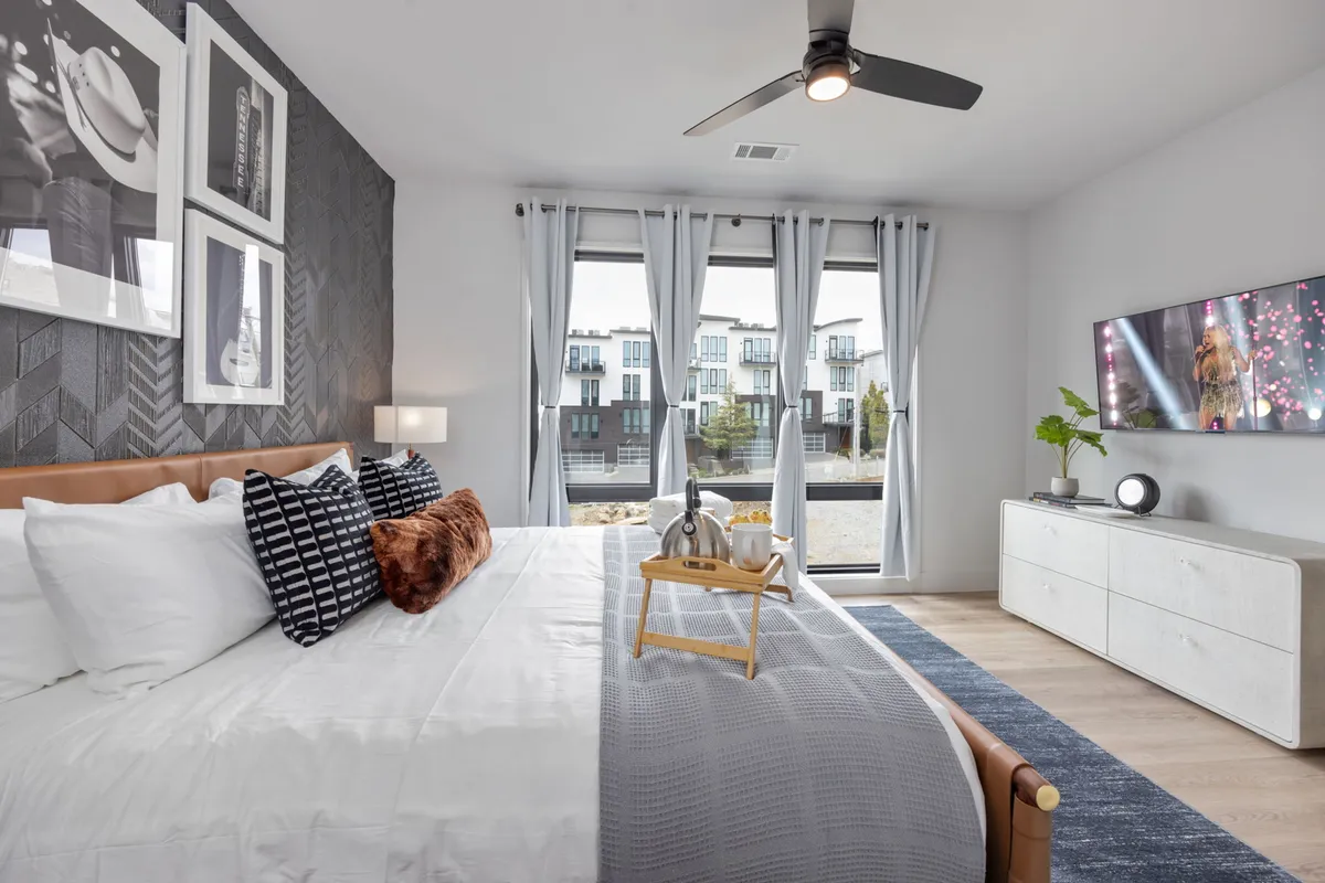 A stylish bedroom featuring a comfortable bed with decorative pillows, large windows draped with curtains for natural light, a wall-mounted television, and tasteful wall art. The overall mood is inviting and restful, perfect for unwinding at the end of the day.