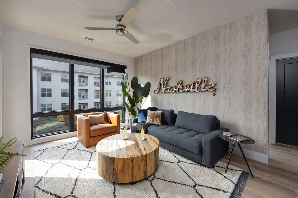A cozy and stylish living area with a large sofa, wooden coffee table, decorative green plant, and a prominent 'Nashville' sign on the wall. Large windows flood the space with sunlight, and the room exudes local character and comfort.