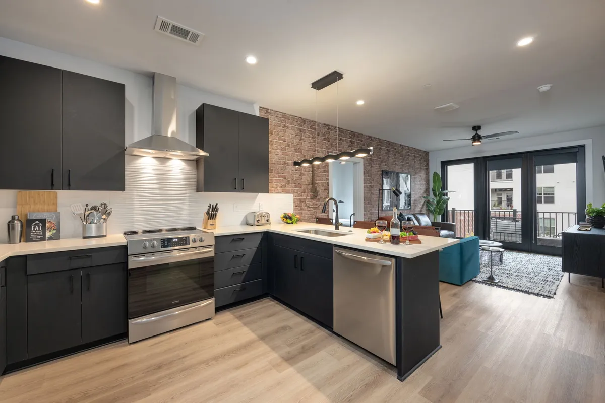 An open-plan kitchen and dining area with a large kitchen island, stainless steel appliances, brick accent wall, and an adjacent dining table. Large windows bring in abundant light, while the space is clearly set up for both cooking and enjoying meals together.